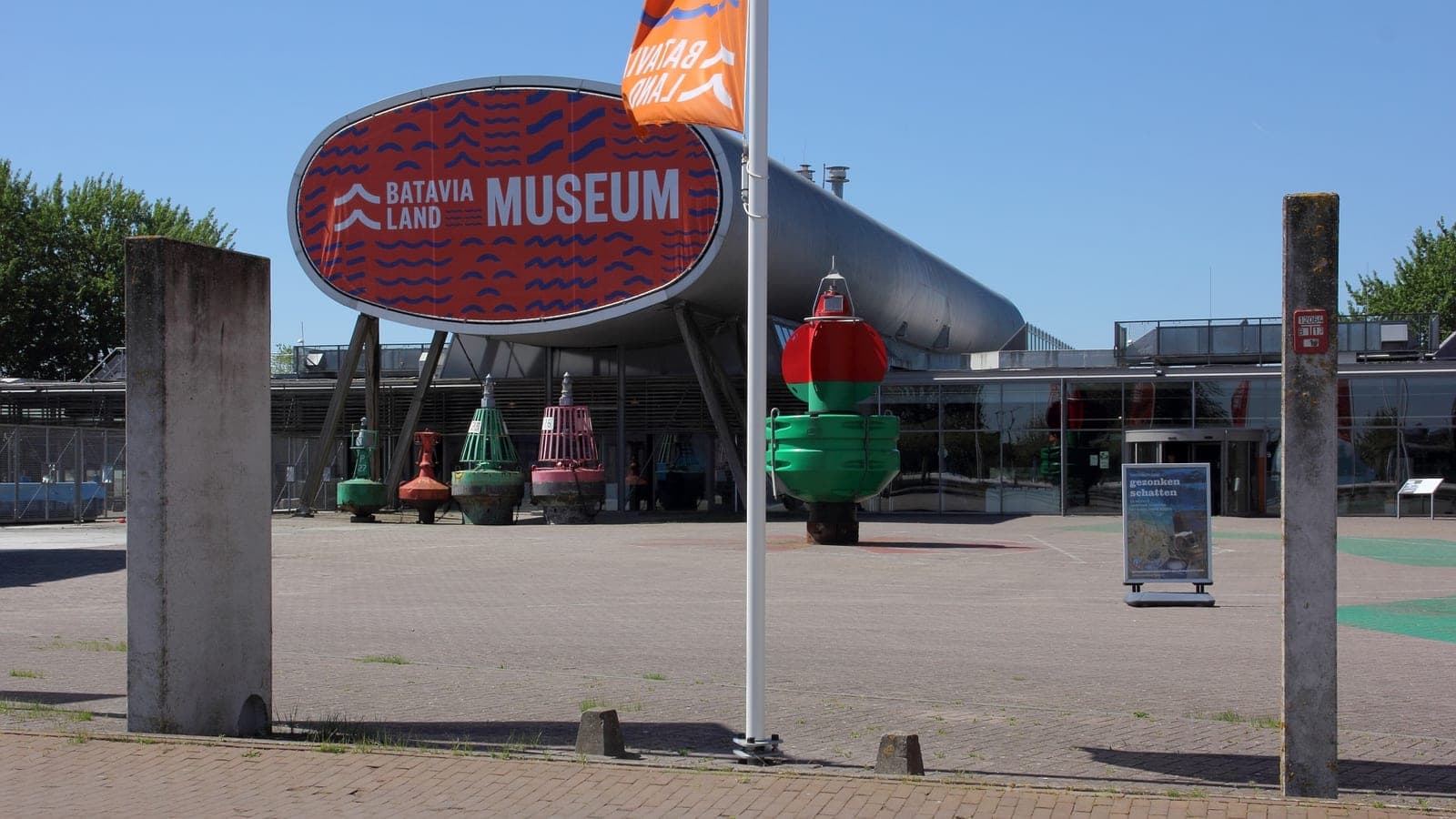 De ingang van Museum Batavialand met het oranje bord en oude boeien op het plein